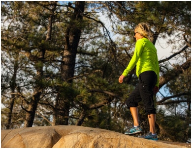 Woman walks on large boulders with tall trees in background