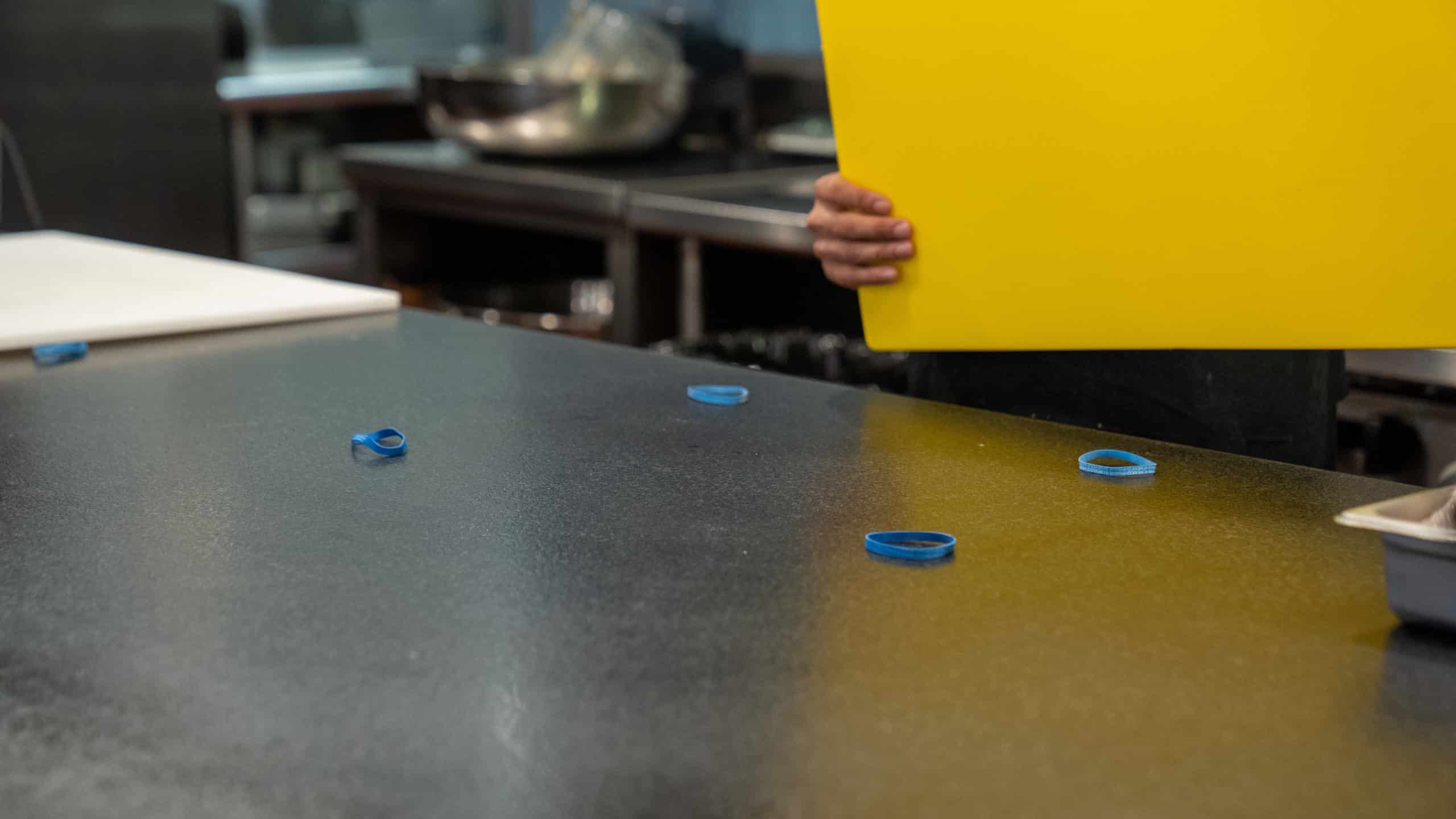 person holds yellow cutting board above four blue rubber bands sitting on gray kitchen counter
