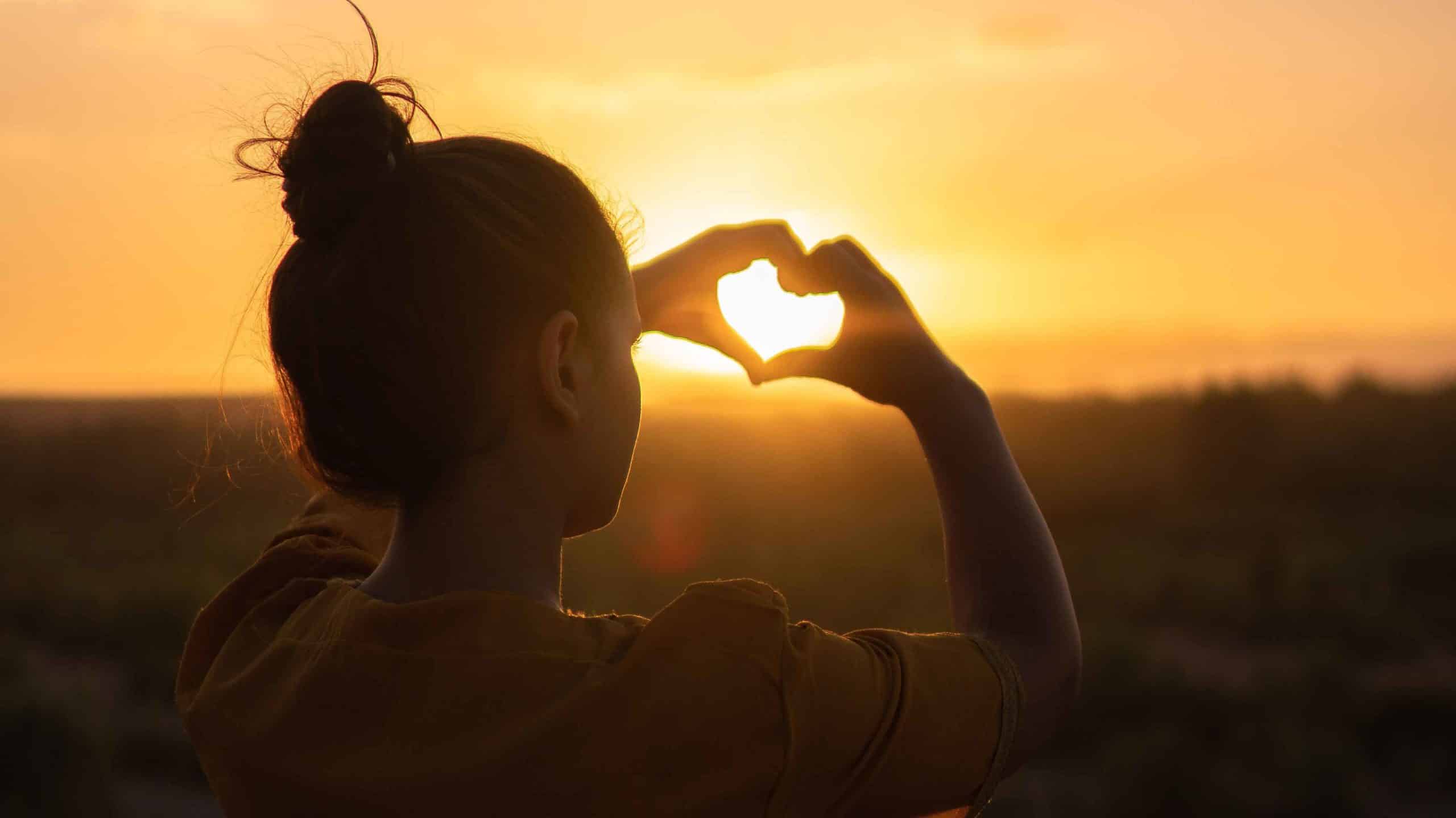 woman makes heart shape with hands against sunset