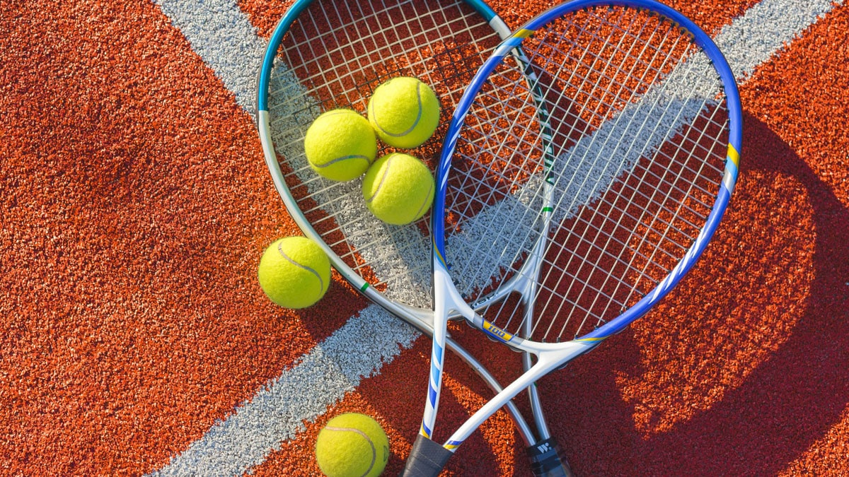 two tennis rackets overlap on an orange tennis court surrounded by green tennis balls